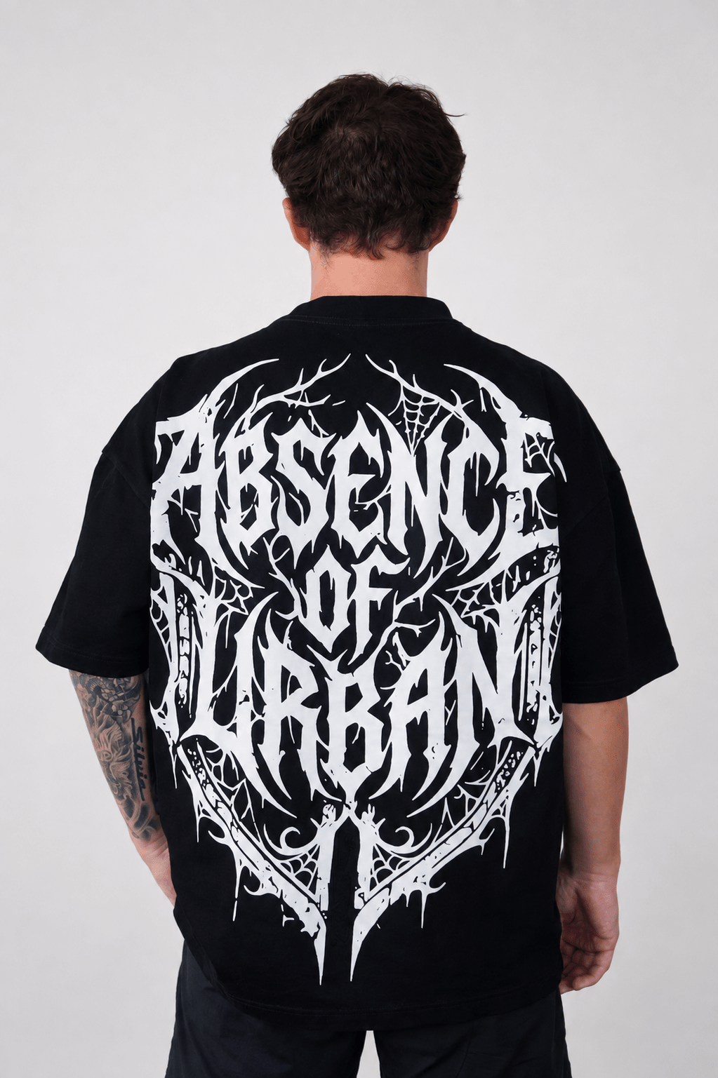 A man viewed from behind wearing a black short-sleeve t-shirt with a large, intricate white gothic-style graphic reading 'ABSENCE OF URBAN' across the back.