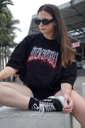 A woman sitting on a concrete ledge outdoors, wearing a black oversized graphic T-shirt with a stylized white and red text design, black shorts, black socks, and black low-top sneakers.