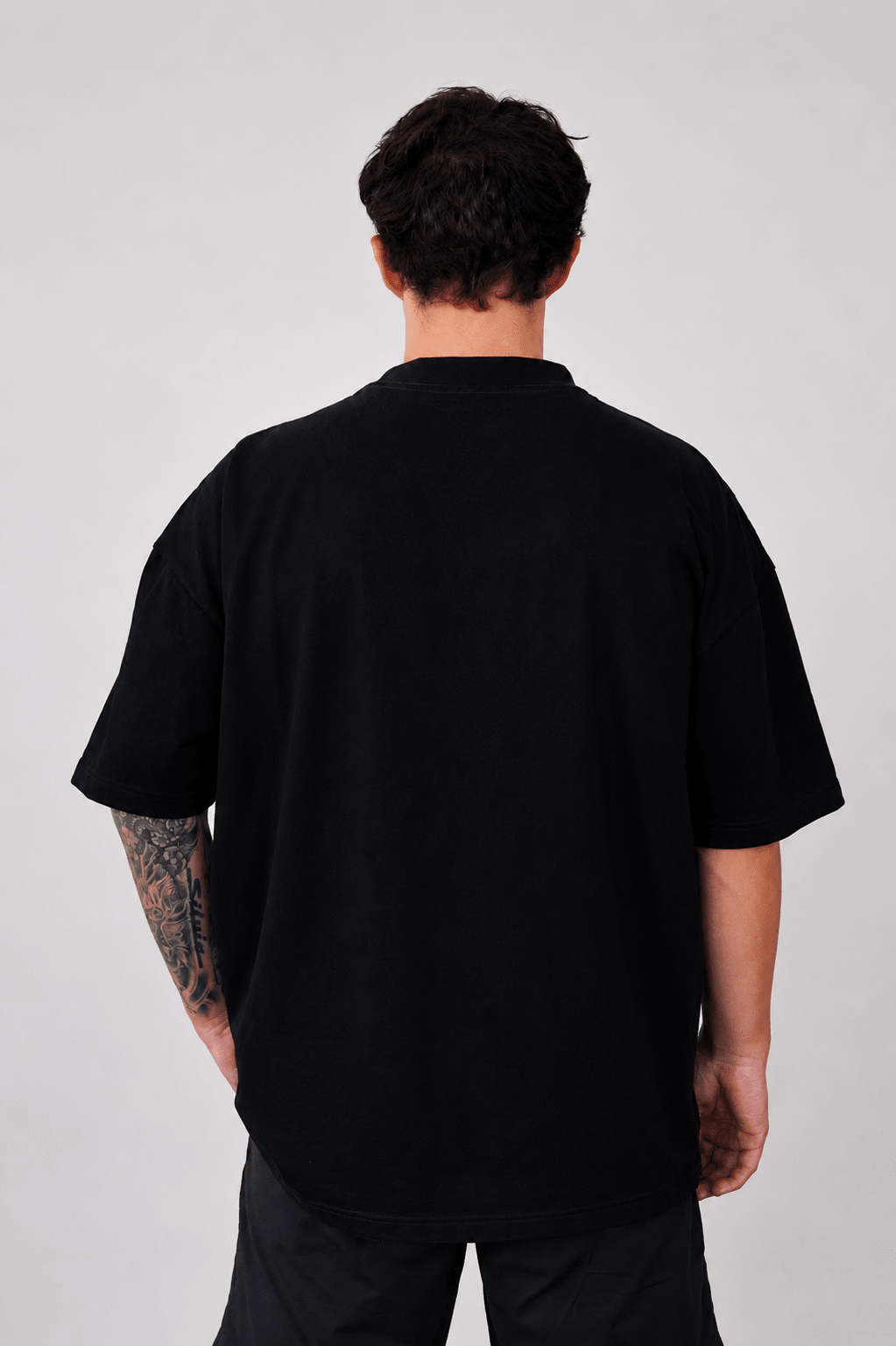 A man viewed from behind wearing an oversized black short-sleeve t-shirt and black pants, standing against a light gray studio background.