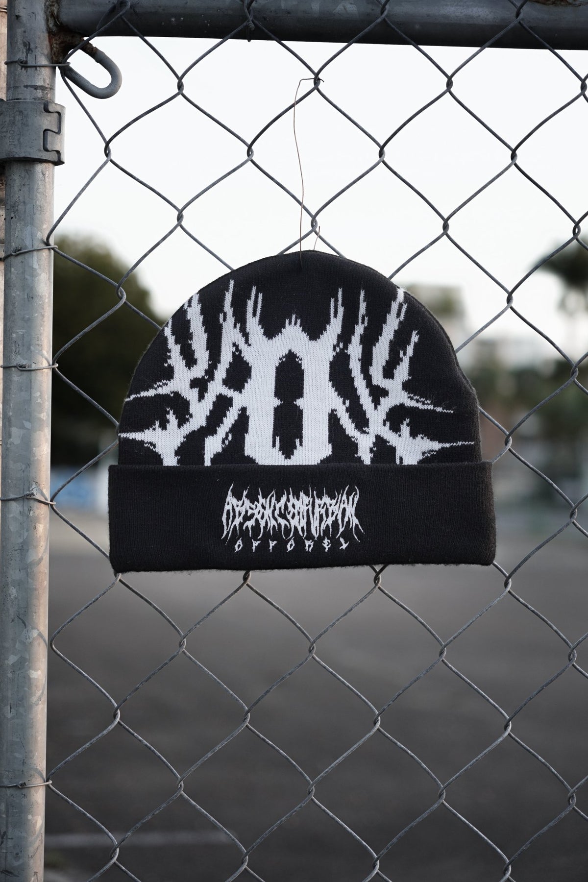 A black knit beanie with a white antler-like graphic and the word 'DEADLY DEATH' in a stylized font, hanging in front of a chain-link fence outdoors.