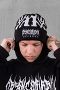 A person wearing a black knit beanie with a white graphic logo and matching black hoodie against a gray concrete wall.
