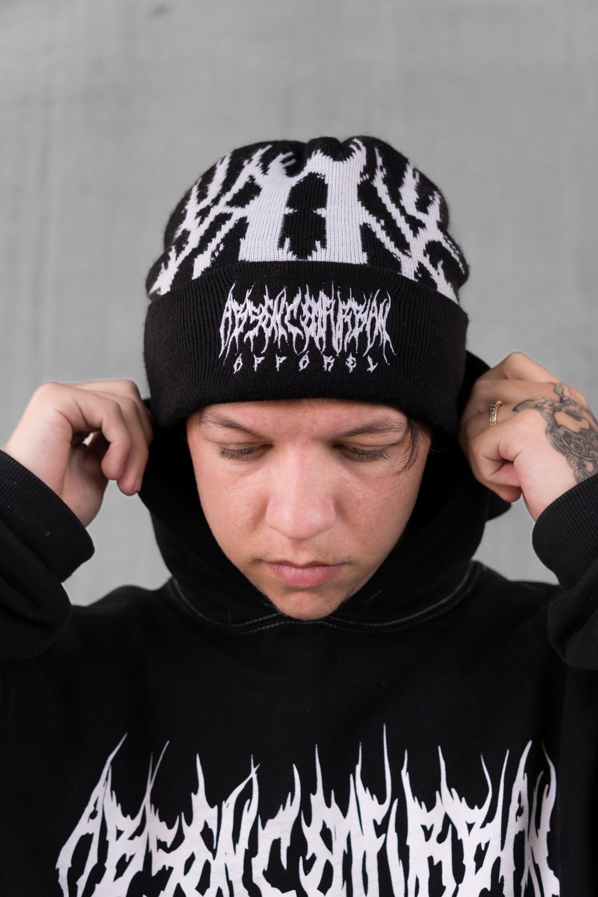 A person wearing a black knit beanie with a white graphic logo and matching black hoodie against a gray concrete wall.
